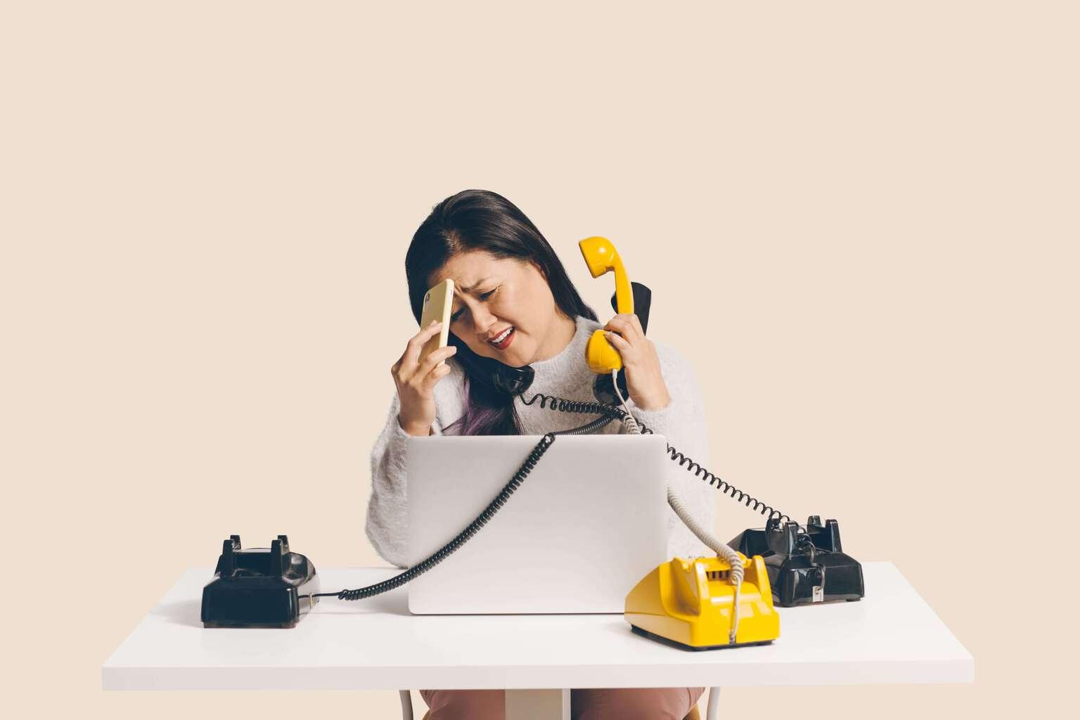 Frau, die an einem Schreibtisch mit mehreren schnurgebundenen Telefonen arbeitet und versucht, mehrere Anrufe gleichzeitig zu beantworten