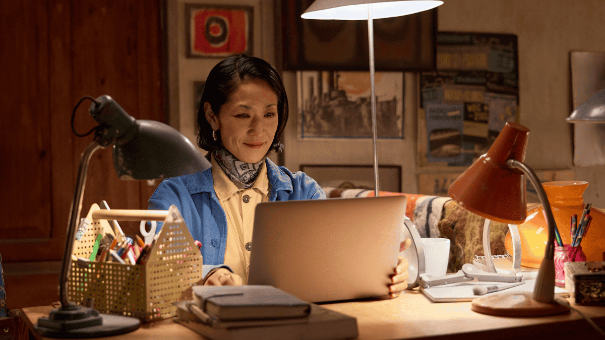 A woman sitting at a wooden desk uses a laptop, surrounded by lamps, books, and stationery.