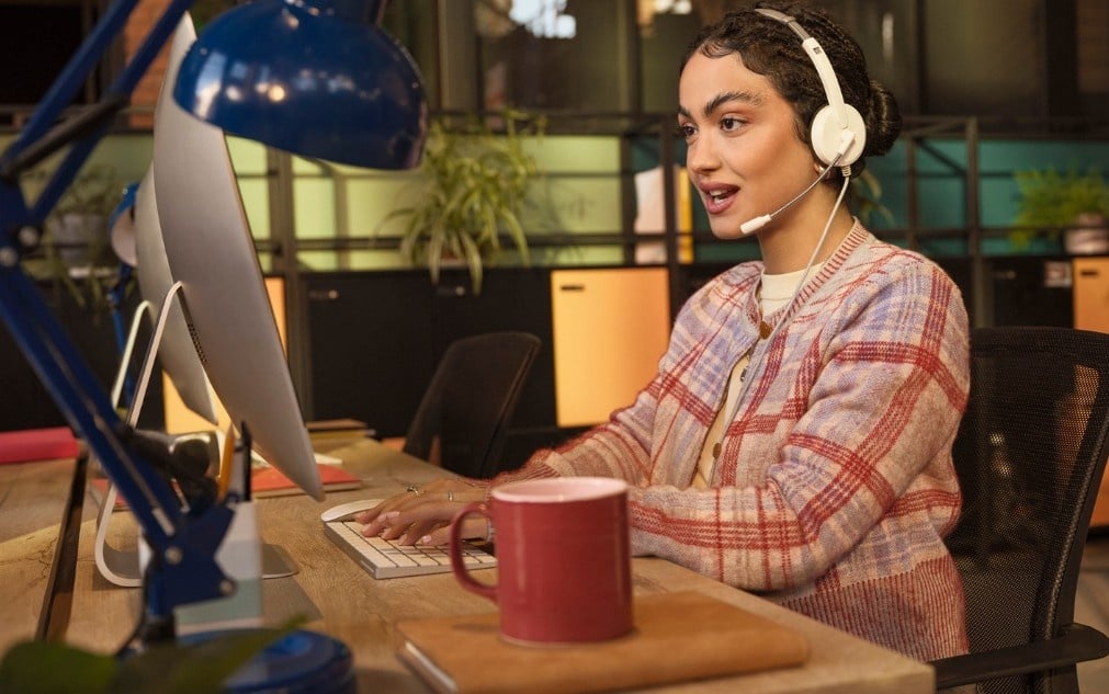 A woman wearing a headset sits at a desk, typing on a computer with a red mug nearby.