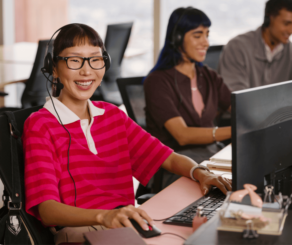 A smiling agent in a pink striped shirt and headset sits at a computer.