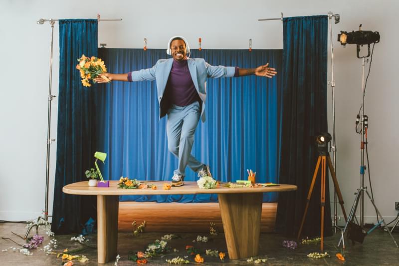 Kundendienst-Mitarbeiter mit Blumenstrauß auf Tisch wie auf Bühne, umgeben von Blumen, Vorhang, Scheinwerfer und Kamera.