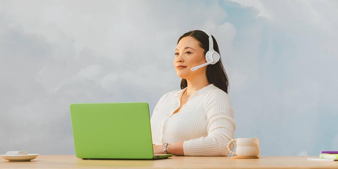 A person wearing a headset, seated at a desk with a green laptop and coffee cup