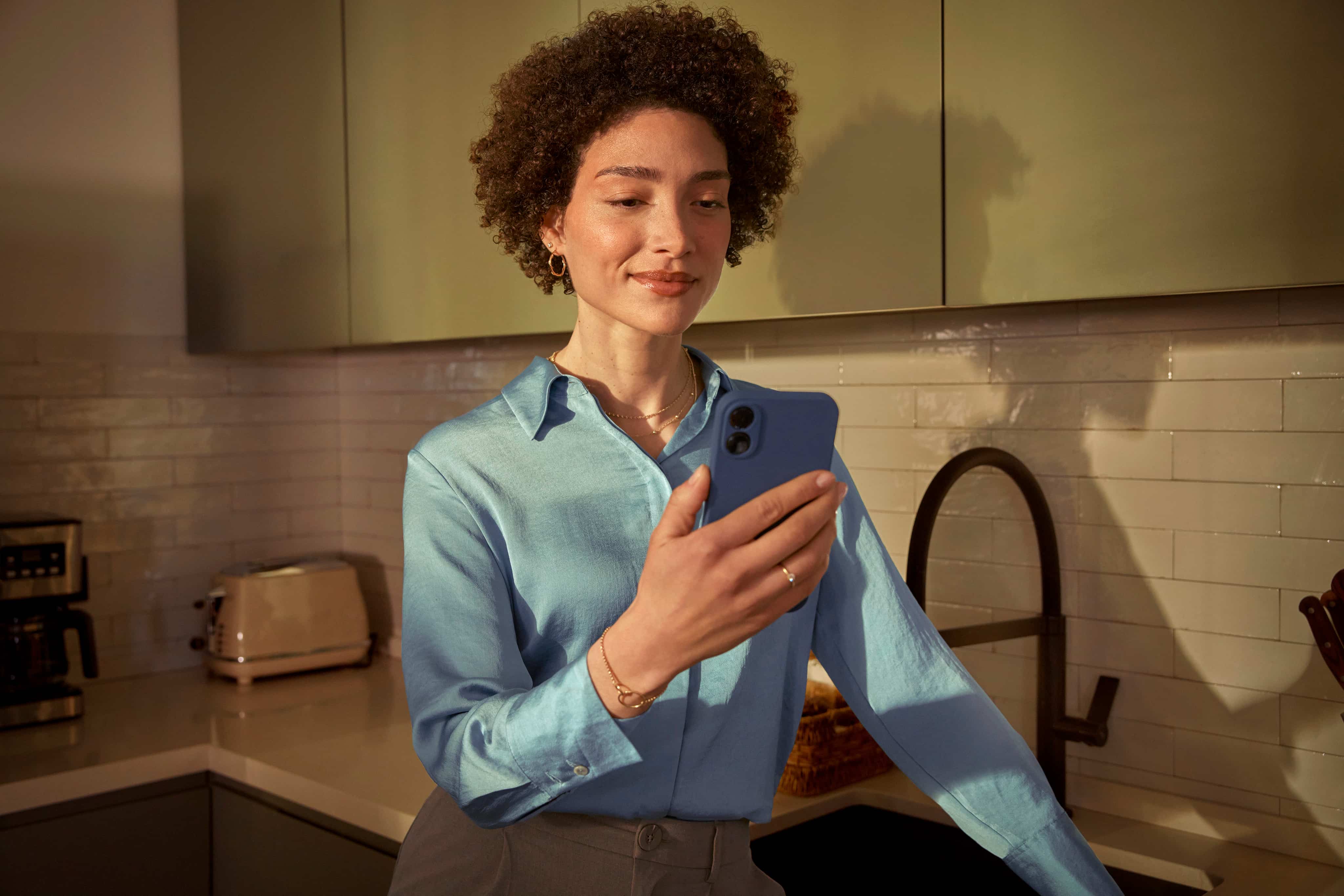 Une femme aux cheveux courts est debout, appuyée contre un comptoir, et regarde son téléphone portable grâce auquel elle reçoit un service client via une IA conversationnelle. 